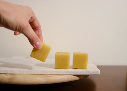 Hand holding a square beeswax candle above two other square beeswax candles on a marble surface.